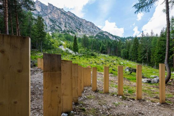 Closer view of tall wooden boards planted in the ground in the middle of a mountain path with high mountains landscape in the background. Sculpture installation by Dado.