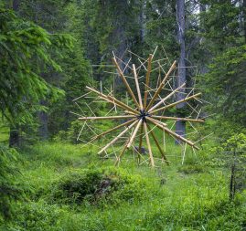 A big wood sculpture composed of clear wood sticks joined together to form a geometrical spherical structure resembling a dandelion, located in a grass field in the middle of a forest, . Artwork by Dado