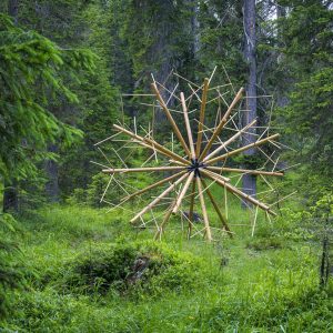 A big wood sculpture composed of clear wood sticks joined together to form a geometrical spherical structure resembling a dandelion, located in a grass field in the middle of a forest, . Artwork by Dado