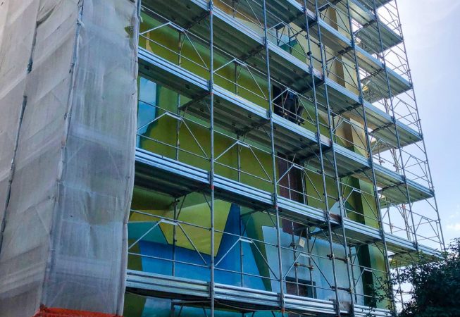 Vista dal basso della facciata del palazzo sulla quale è montata l'impalcatura per la realizzazione del dipinto murale Il Valore della Casa dell'artista Dado