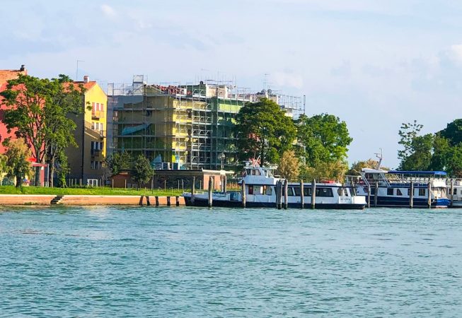 Vista panoramica di uno scorcio di laguna veneziana dove al di la delo specchio d'acqua si vedono, in lontananza, due barche attraccate e, poco più distante, il palazzo sul quale è montata l'impalcatura per la realizzazione del dipinto murale Il Valore della Casa dell'artista Dado