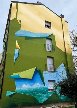 Vista frontale della facciata del palazzo sulla quale realizzato il dipinto murale Il Valore della Casa dell'artista Dado. Il palazzo sembra essere stato "scartato" dal suo involucro verde pisello e nella parte alta svela il vero colore dorato del palazzo.