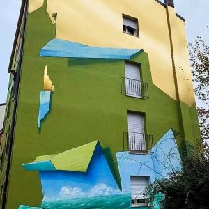 Vista frontale della facciata del palazzo sulla quale realizzato il dipinto murale Il Valore della Casa dell'artista Dado. Il palazzo sembra essere stato "scartato" dal suo involucro verde pisello e nella parte alta svela il vero colore dorato del palazzo.