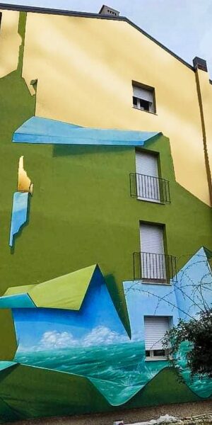 Vista frontale della facciata del palazzo sulla quale realizzato il dipinto murale Il Valore della Casa dell'artista Dado. Il palazzo sembra essere stato "scartato" dal suo involucro verde pisello e nella parte alta svela il vero colore dorato del palazzo.