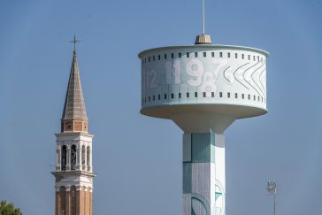 Vista da lontano della parte superiore della del grande serbatoio idrico di Mira, provincia di Venezia, sul quale è dipinto l'Orologio Solare senza Gnomone dell'artista Dado. Dietro alla testa del grande serbatoio, vista quasi frontalmente, è visibile la cima di un campanile, della stessa altezza, in stile rinascimentale,