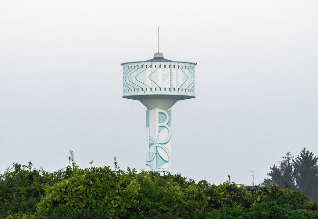 Vista da lontano della parte superiore della del grande serbatoio idrico di Mira, provincia di Venezia, sul quale è dipinto l'Orologio Solare senza Gnomone dell'artista Dado. La testa del grande serbatoio, vista quasi frontalmente, è stagliata sul un pallido cielo bianco.